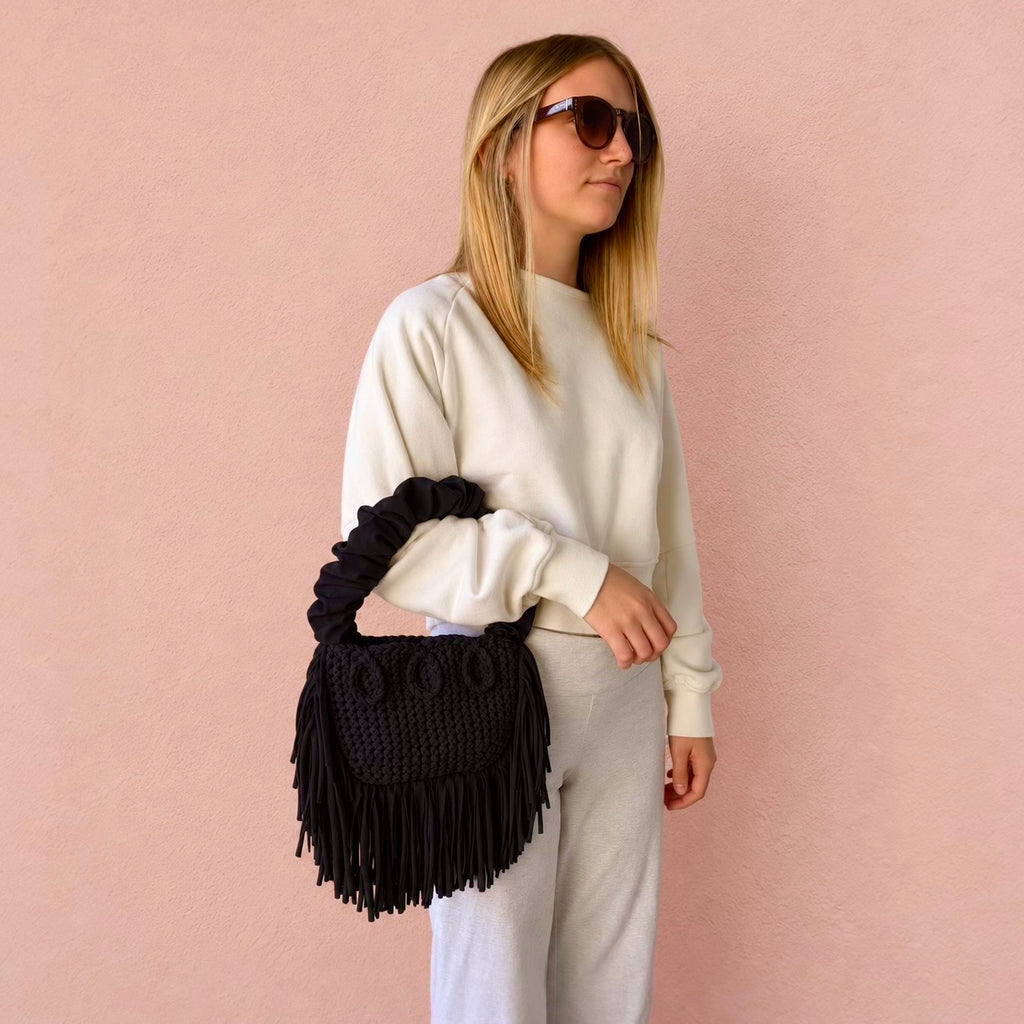 Onyx black handmade crochet Cha Cha Mini bag featuring a crescent-shaped body, braided loop detailing, ruched scrunchie handle and flowing fringe. 